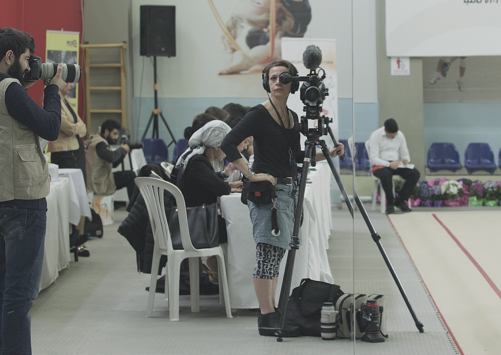 Filmmaker operating a video camera on a tripod while another photographer works nearby at an indoor event.