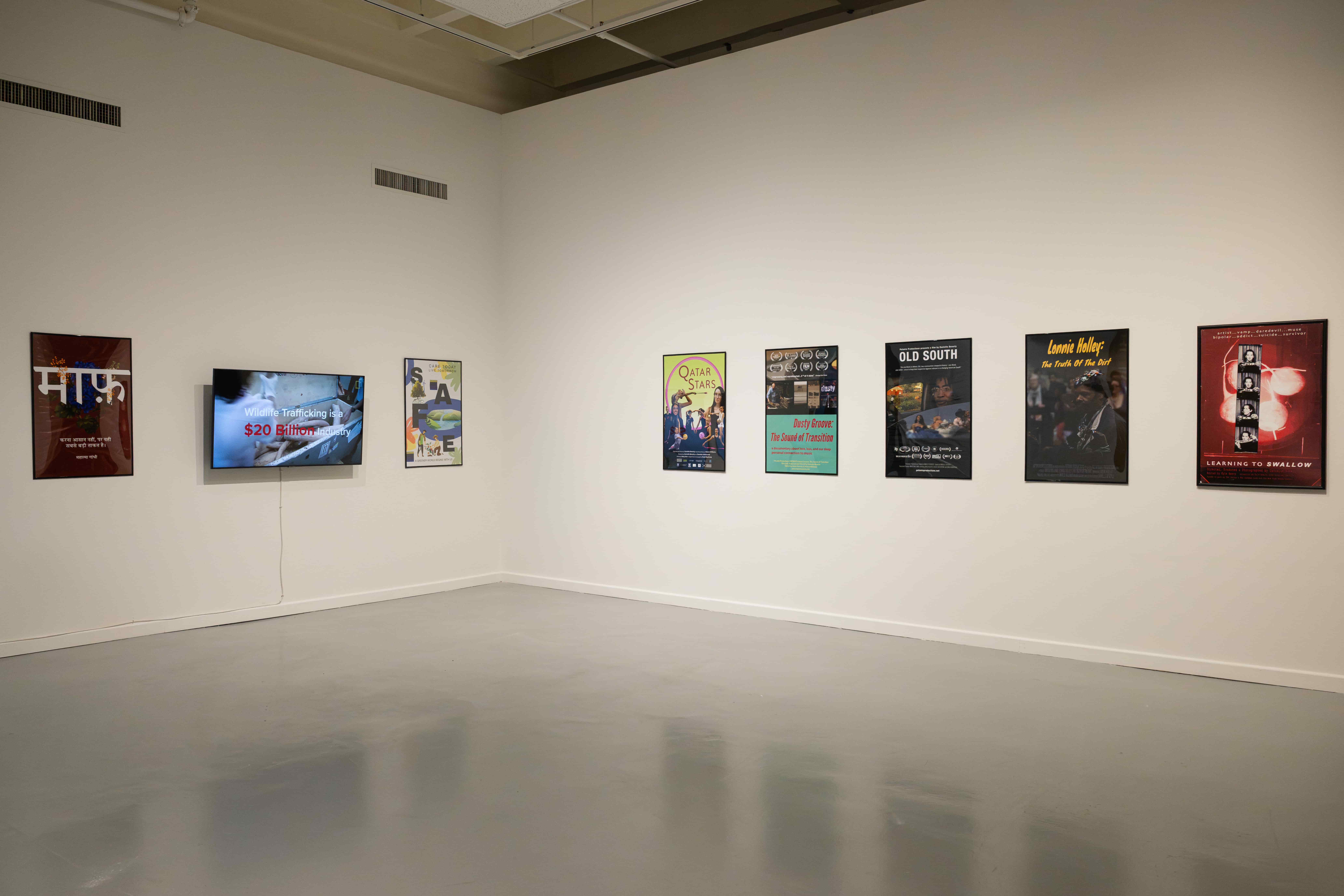 A quiet art gallery corner displays colorful graphic posters along white walls above a bare floor, creating a calm, minimalist exhibition mood.