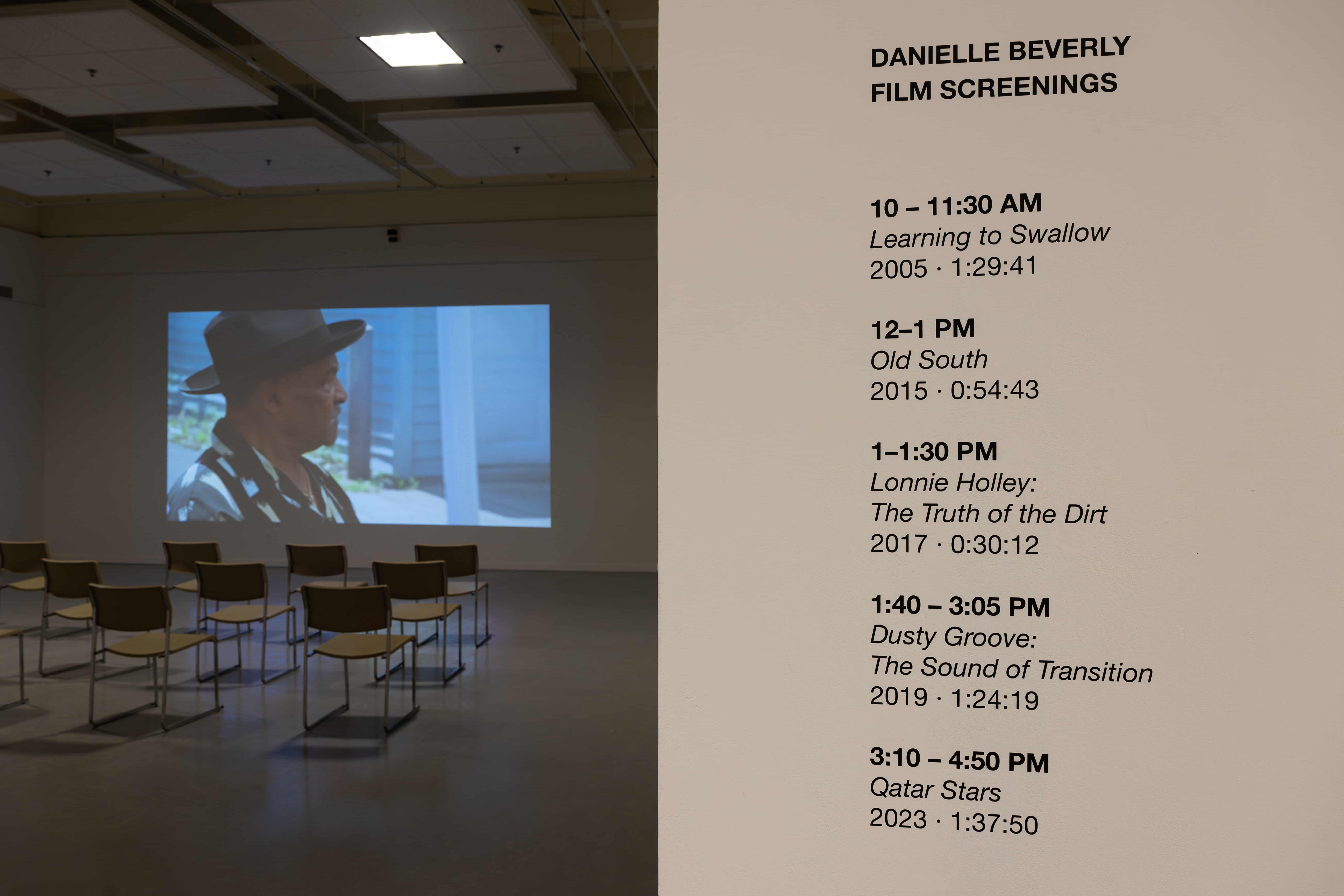 A gallery screening room shows a projected film still of a man in a hat, with empty chairs facing the screen; a wall lists Danielle Beverly film screenings.