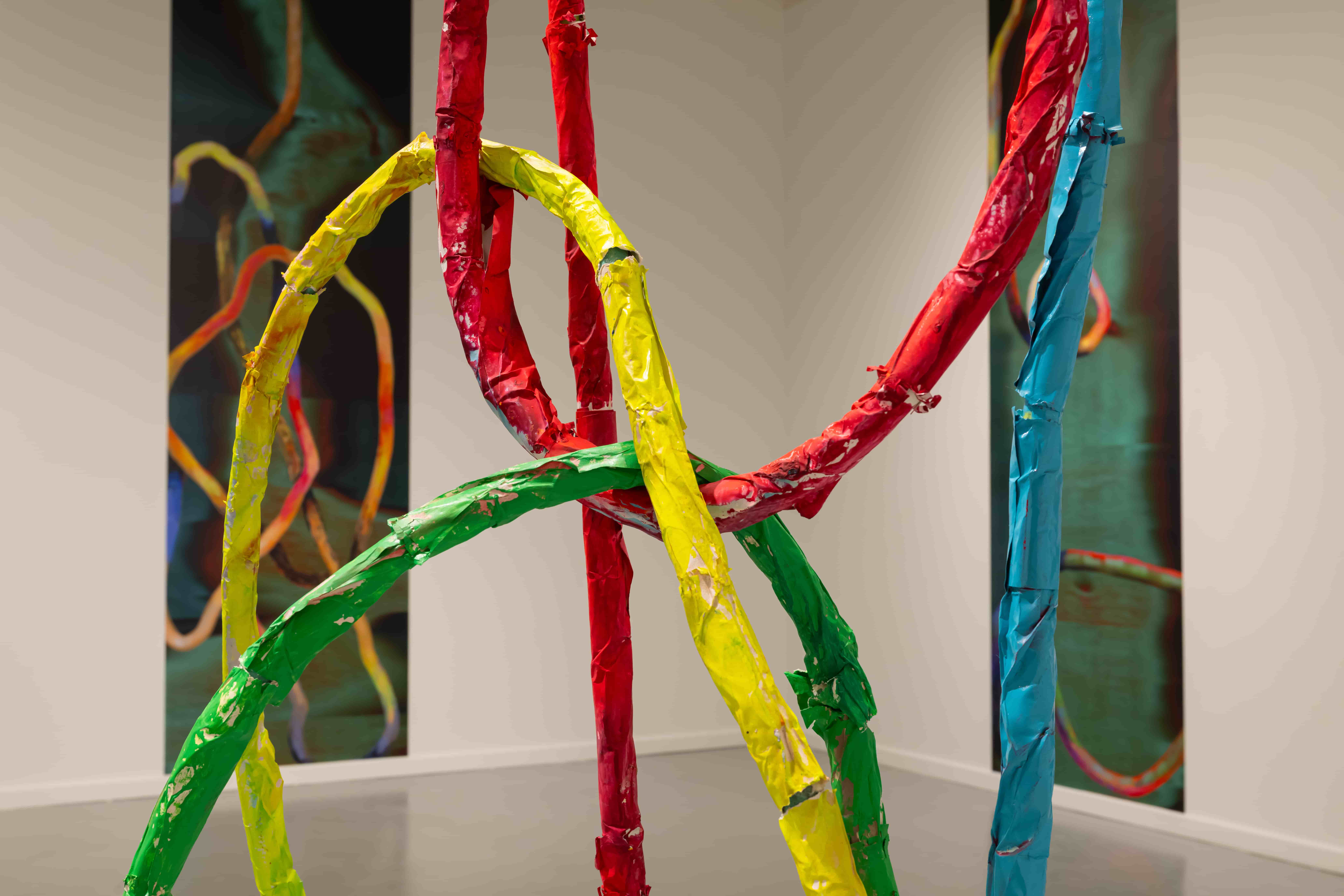 Colorful abstract sculpture of looping red, yellow, green, and blue strips in a gallery, creating a playful, tangled sense of movement.