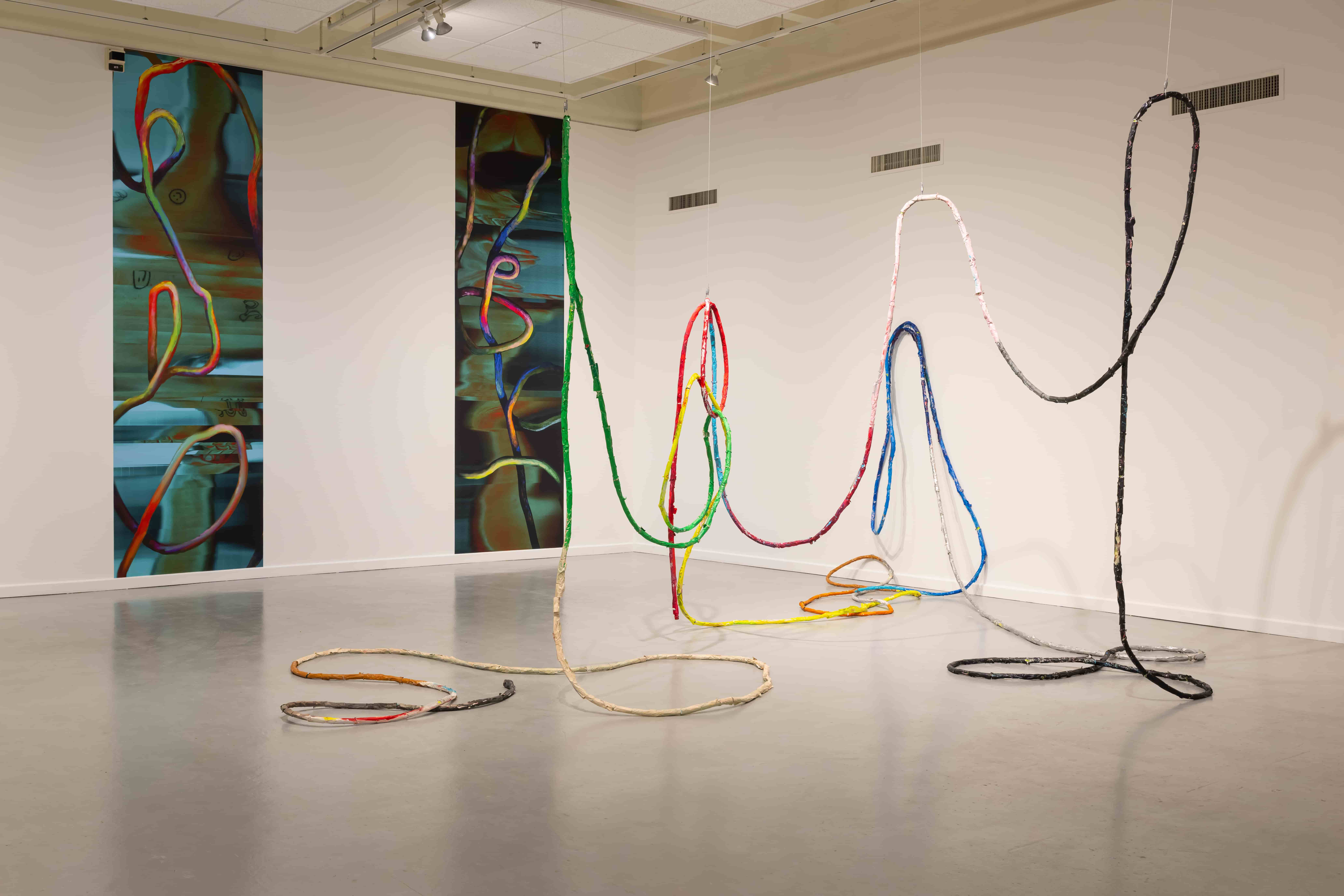A contemporary art installation features colorful looping cables across a gallery floor, echoing abstract paintings on the walls and creating a playful, exploratory mood.