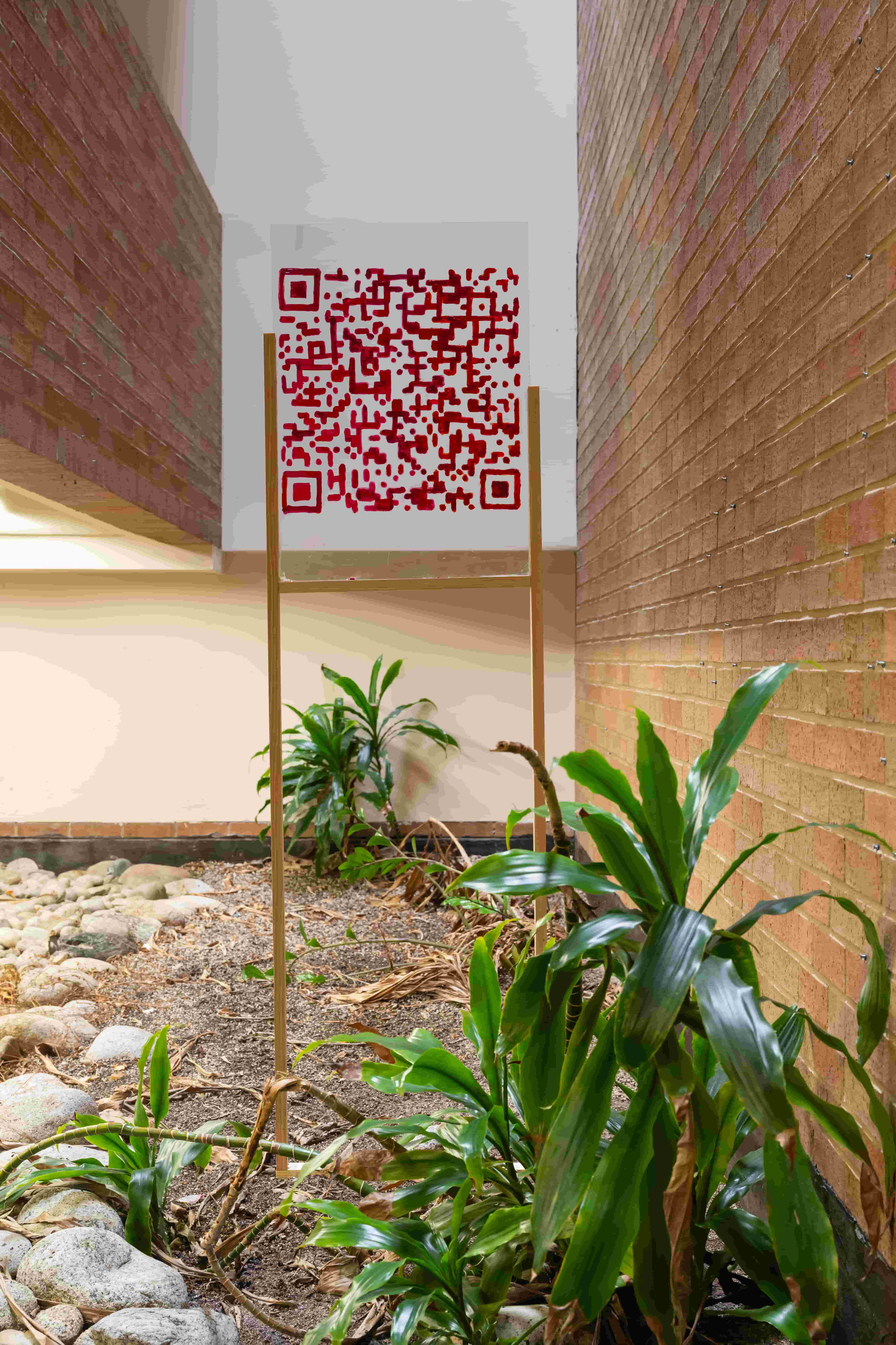 A red QR code sign stands on a slim frame in a narrow brick courtyard, surrounded by green plants and pale stones; the space feels calm and enclosed.